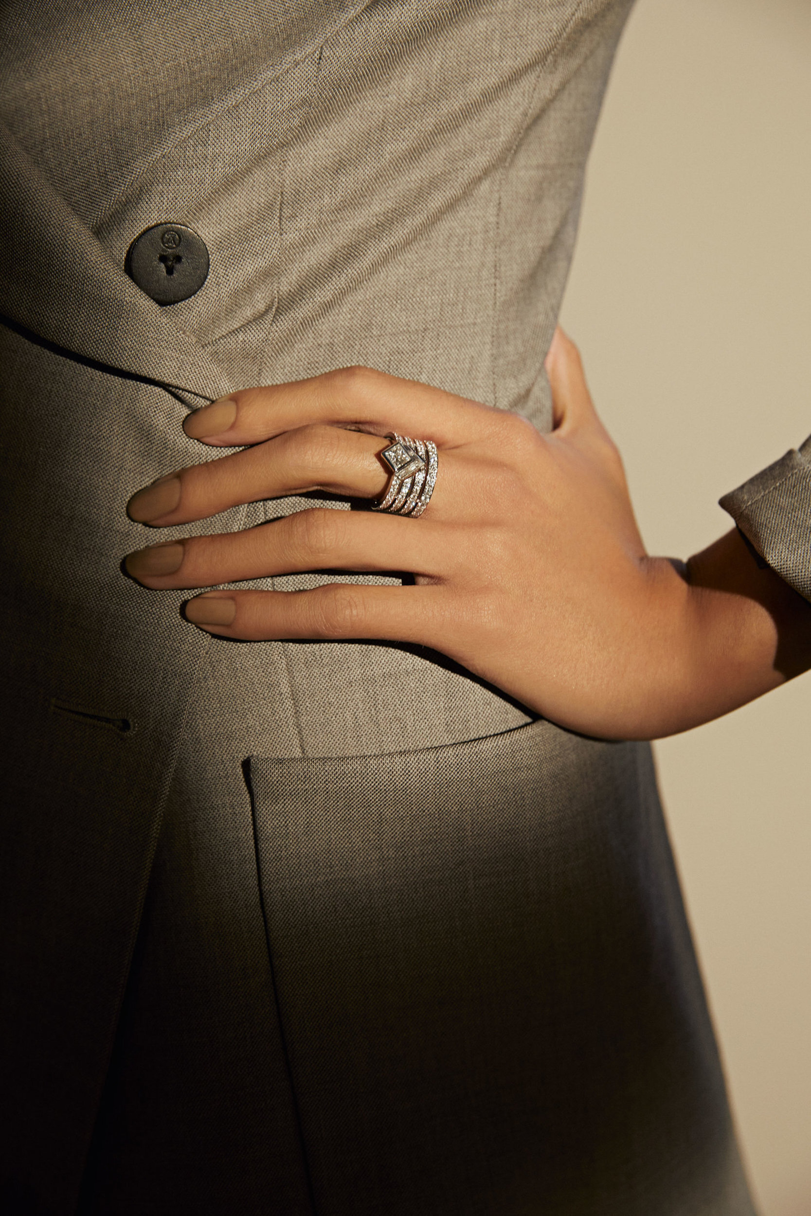 Close-up shot of an adult female's hand adorned with a diamond ring, styled with neutral nail polish, and a grey blazer sleeve.