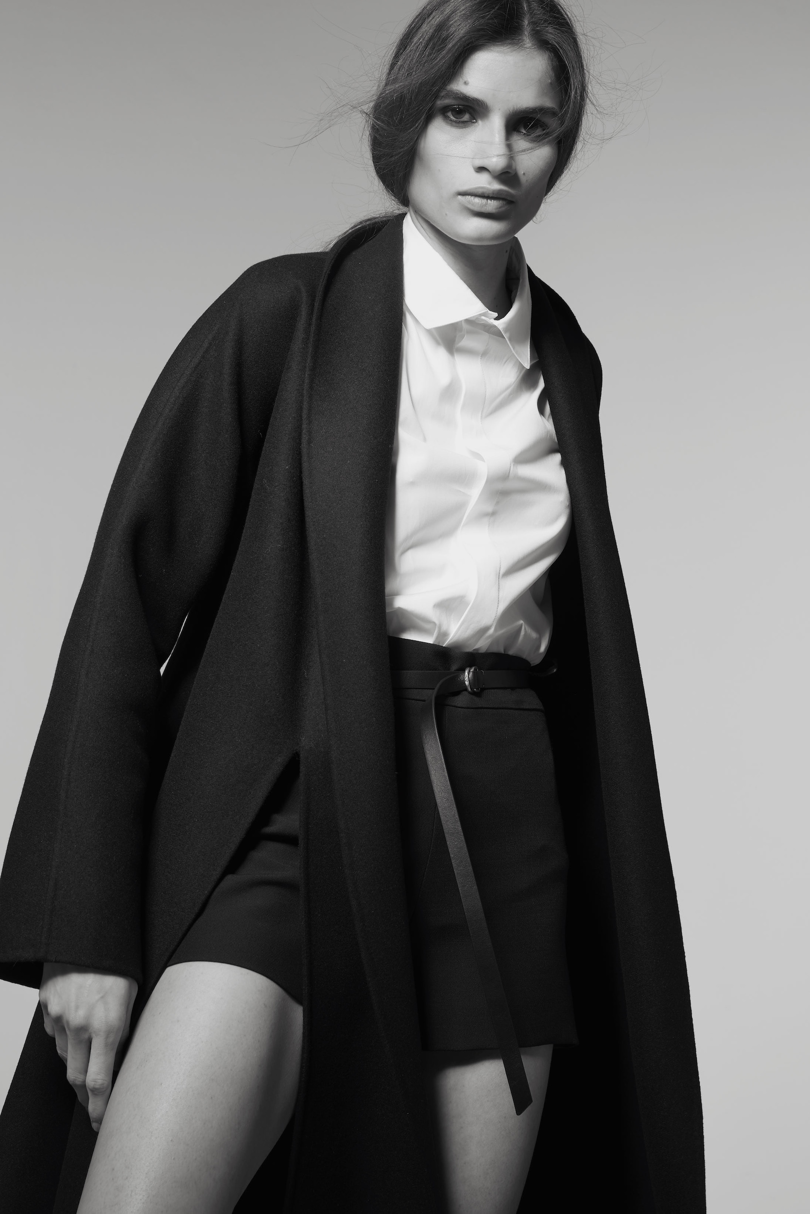 Monochrome studio shot of a woman in a white shirt and black shorts with a wrap coat, gazing directly at the camera.