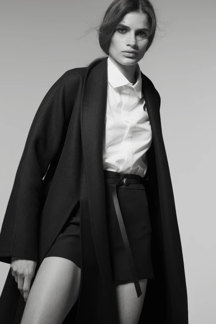Monochrome studio shot of a woman in a white shirt and black shorts with a wrap coat, gazing directly at the camera.