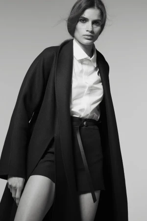 Monochrome studio shot of a woman in a white shirt and black shorts with a wrap coat, gazing directly at the camera.