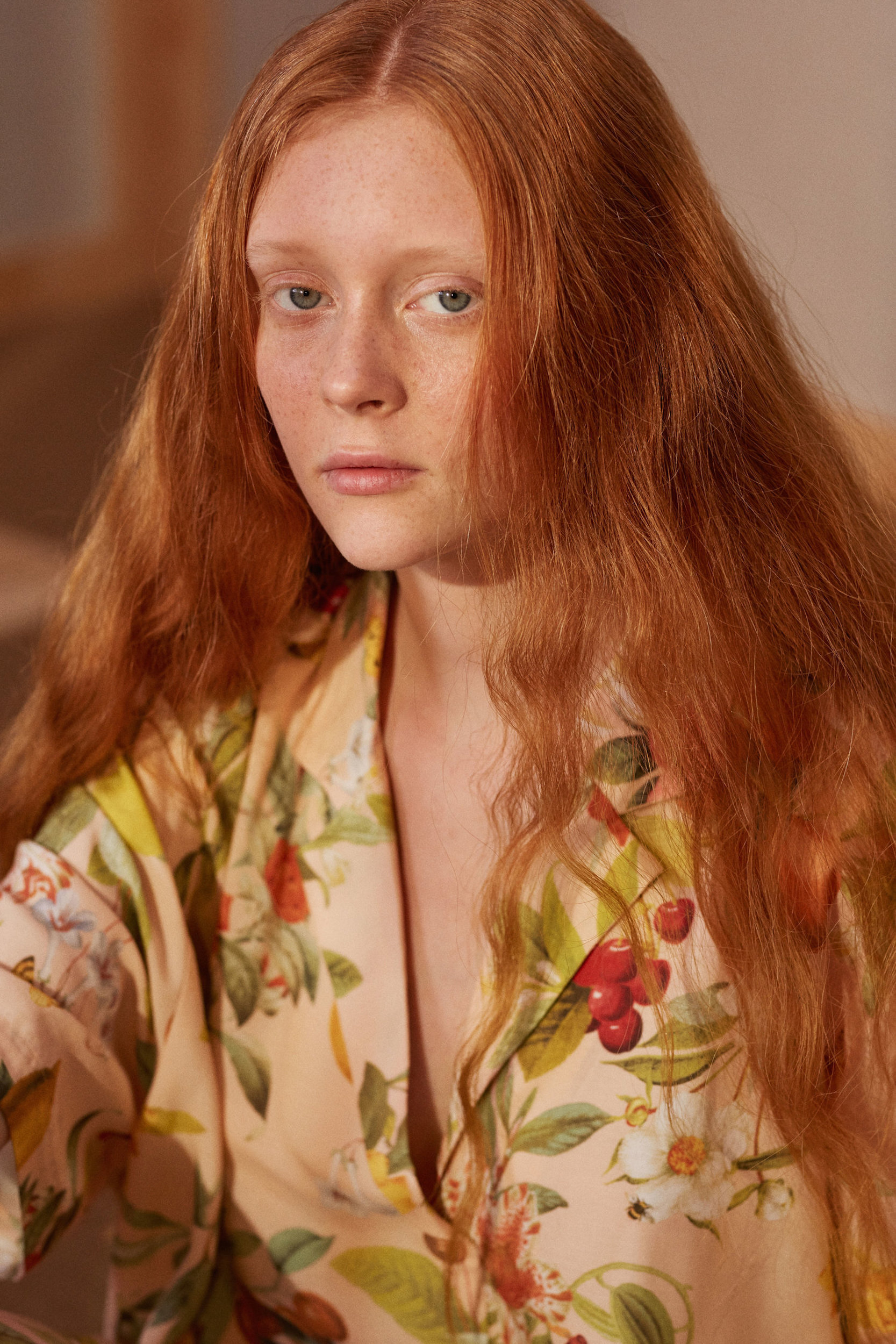 Close-up portrait of a young woman with red hair wearing a floral-patterned shirt looking directly at the camera with a neutral expression.