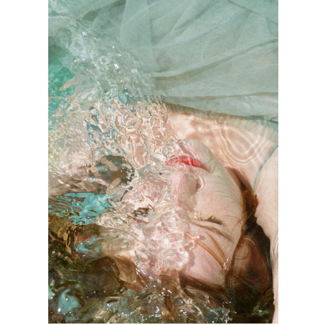 Woman's face emerges from water with rippling surface texture, sheer fabric backdrop, creating a serene and ethereal underwater portrait.