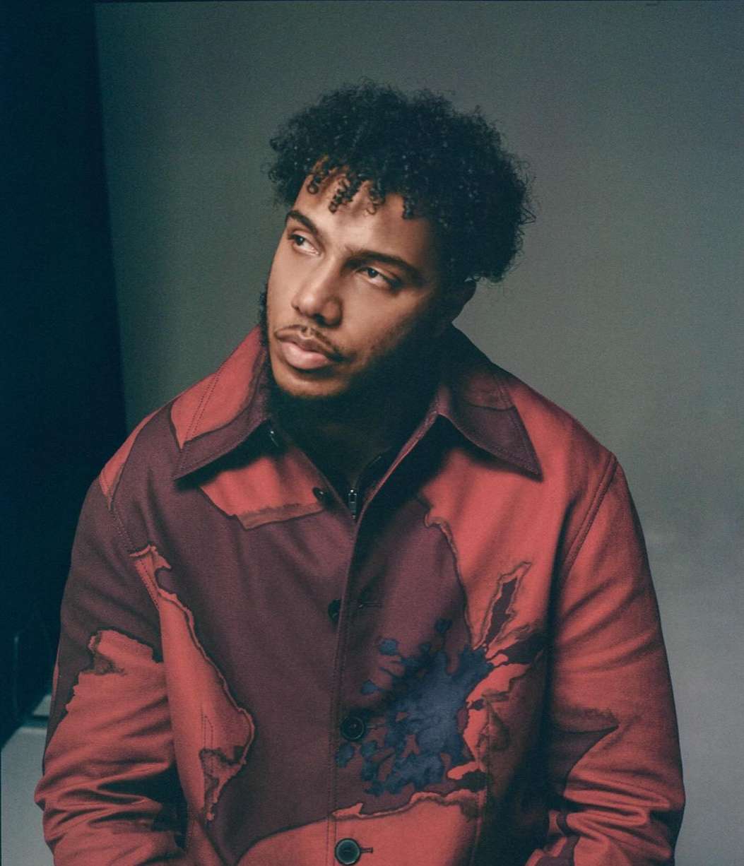 Portrait of a Black man with curly hair wearing a patterned red jacket against a grey studio backdrop.