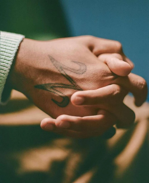 Close-up shot of clasped hands of a person with tan skin, a tattoo visible on the back of the top hand, wearing a beige sweater.