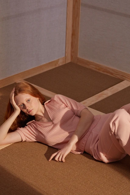 Red-haired adult female wearing a pink matching set lies on a brown geometric patterned floor in a corner with natural light.