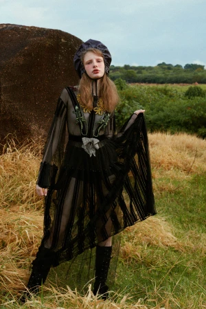 A model poses in a black dress and hat outdoors in a field of tall grass with a cloudy sky, showcasing fashion and nature.
