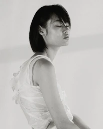 Monochrome portrait of an East Asian woman with eyes closed wearing a white sleeveless top against a plain light background.