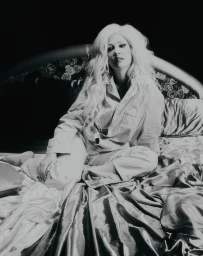 Woman with long blonde hair seated on a glossy bed wearing pajamas, captured in a monochrome bedroom scene.