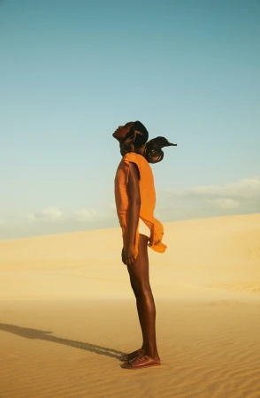 Black woman in orange tunic standing on sand dunes with her head tilted upward under a clear sky.