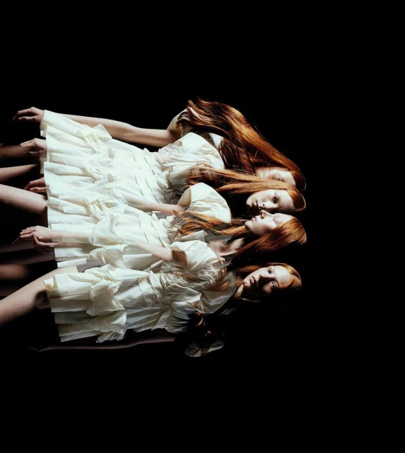 Multiple red-haired women in white dresses lie against a dark backdrop, creating a surreal, almost mirrored effect in a studio setting.