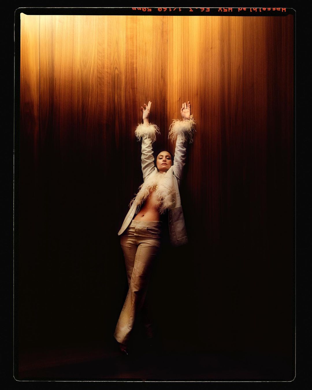 Woman in light-colored suit with feathered trim poses against a wood panel wall in a studio with dramatic lighting.