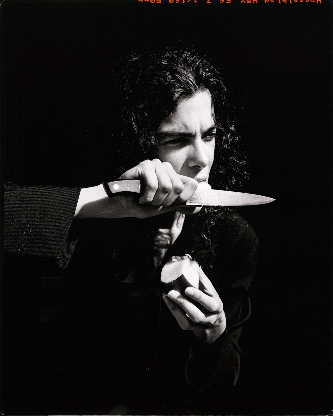 Portrait of a person holding a knife near their face with an apple in hand against a dark background