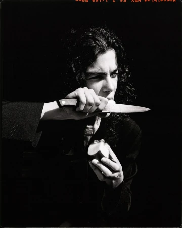 Portrait of a person holding a knife near their face with an apple in hand against a dark background