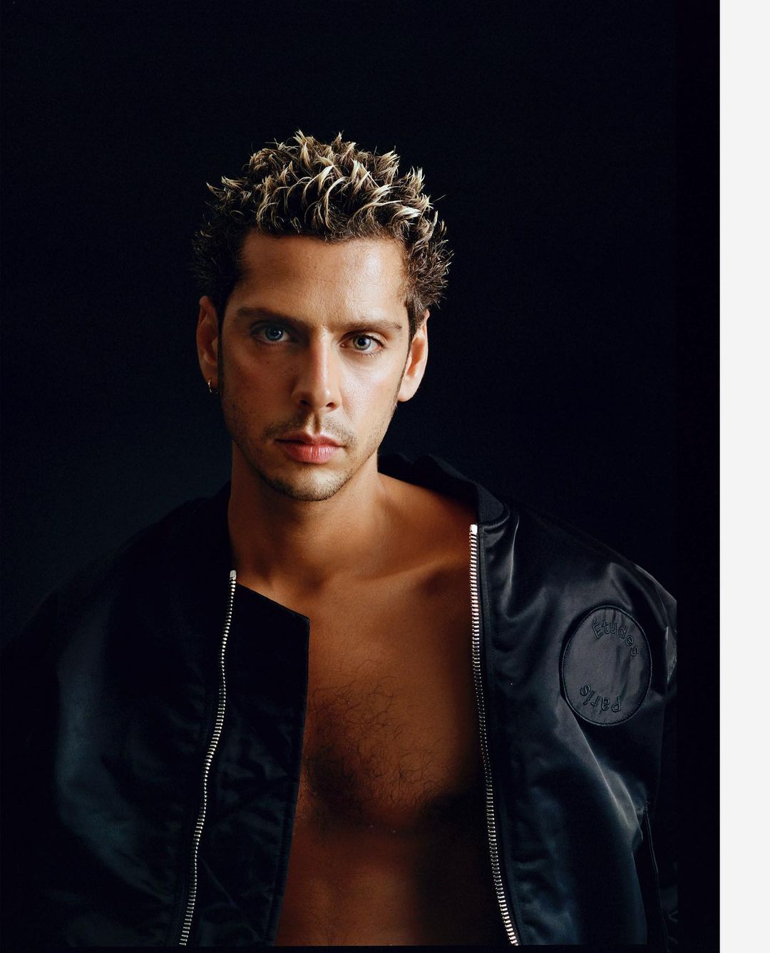 Portrait of a shirtless adult male with blond spiky hair wearing a black jacket, posing against a dark background, eye-level angle.