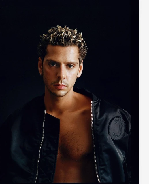 Portrait of a shirtless adult male with blond spiky hair wearing a black jacket, posing against a dark background, eye-level angle.