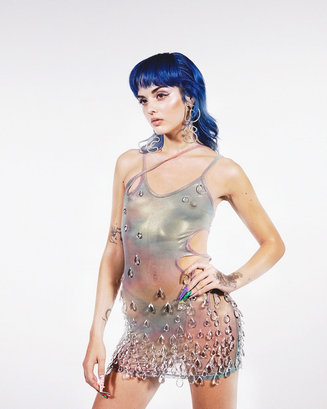 A young adult woman with blue hair poses in a fashion forward iridescent and clear beaded mini dress in a bright photo studio setting.