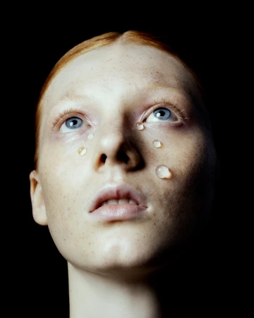 Close-up portrait of a woman with red hair and blue eyes, with water droplets on her face against a black background.