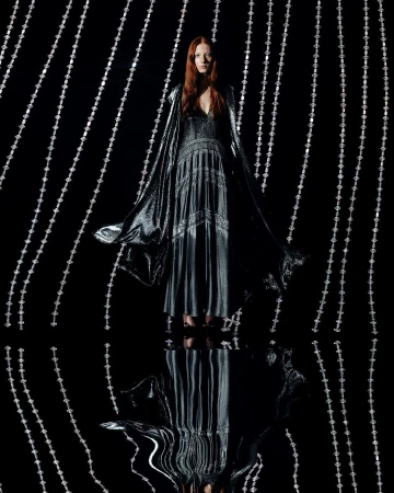 Full-length shot of a woman with red hair in a long dark dress with a cape, standing on a reflective surface with beaded string lights.