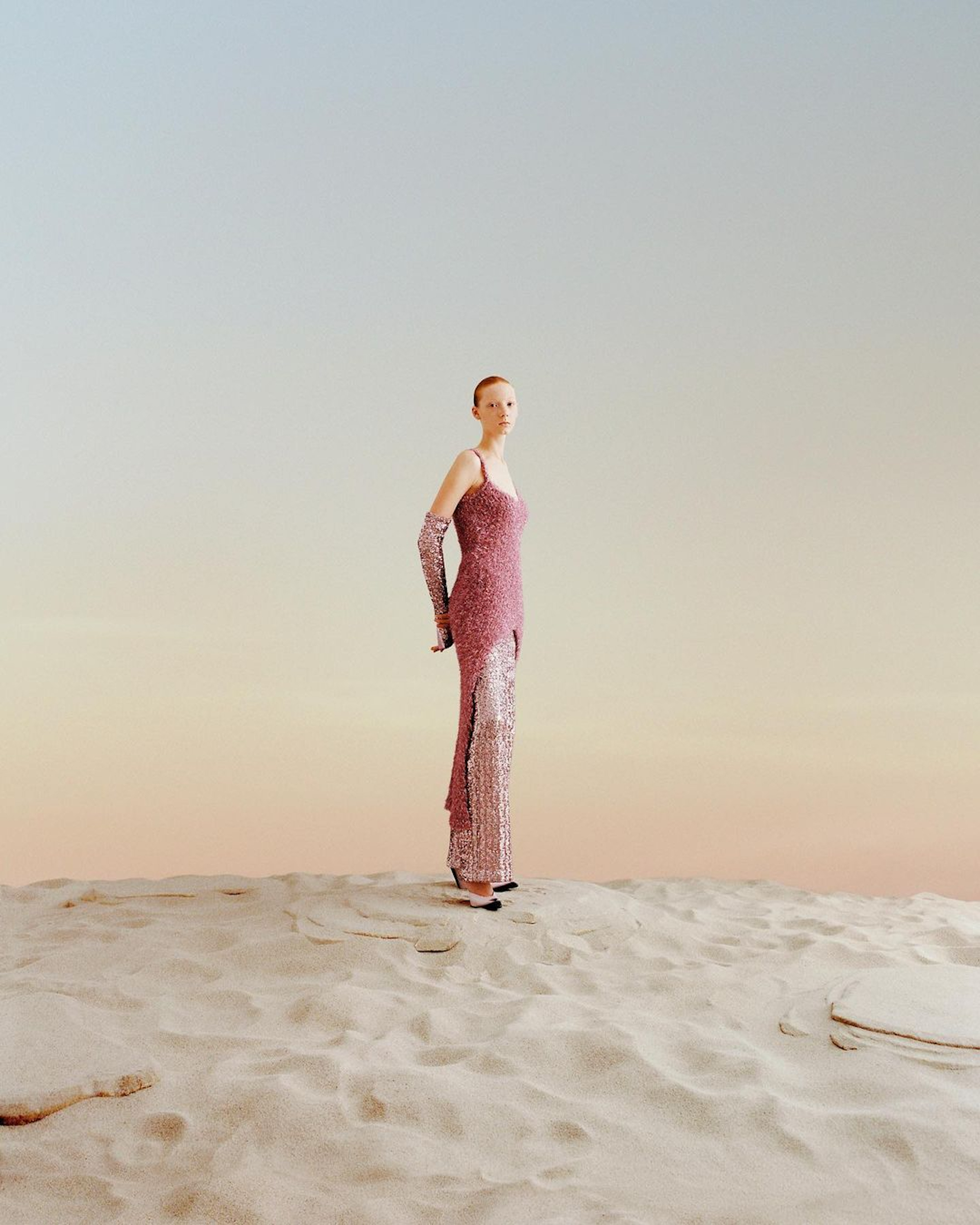 Full shot of a person in a pink sequined dress with matching gloves on sand, set against a light gradient blue to light orange sky.