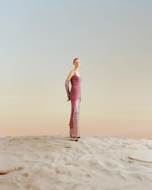 Full shot of a person in a pink sequined dress with matching gloves on sand, set against a light gradient blue to light orange sky.