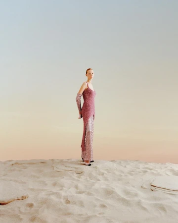 Full shot of a person in a pink sequined dress with matching gloves on sand, set against a light gradient blue to light orange sky.