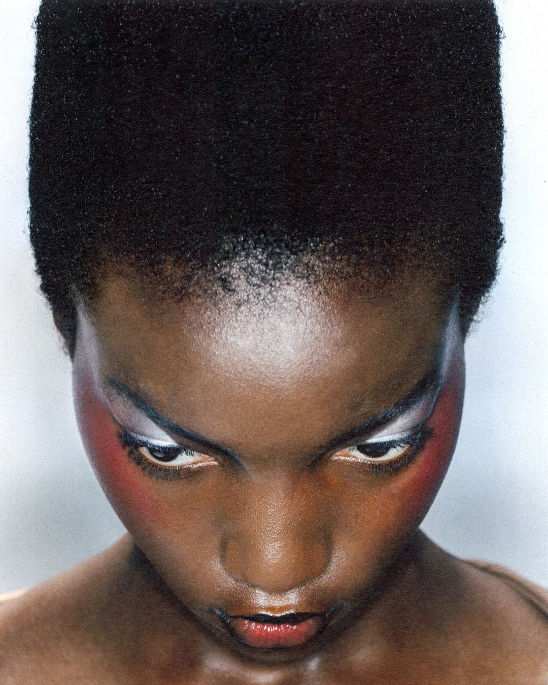 Close-up portrait of an adult female with dark skin, short afro hairstyle, and creative colorful makeup, against a plain white backdrop.