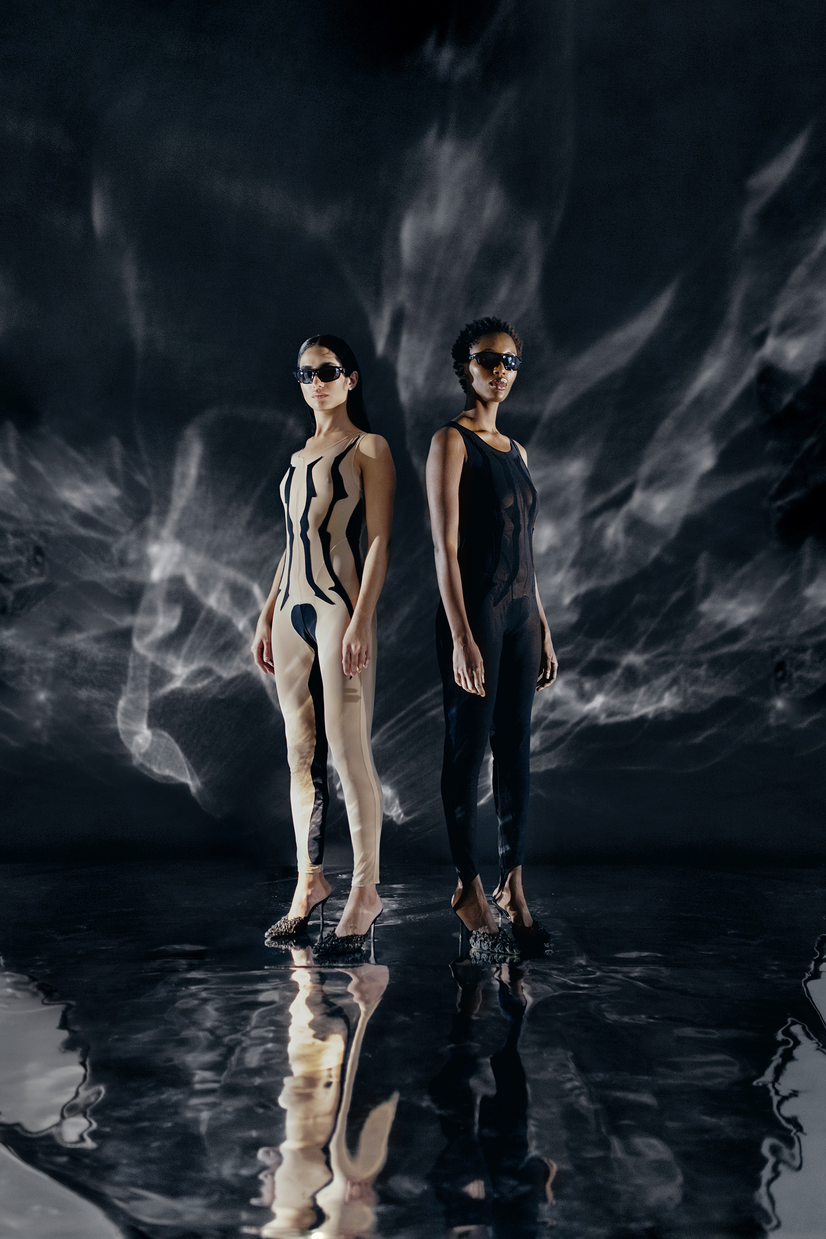 Two women in sleek bodysuits pose against a water-ripple backdrop, showcasing fashion and reflections in a studio setting with dramatic lighting.