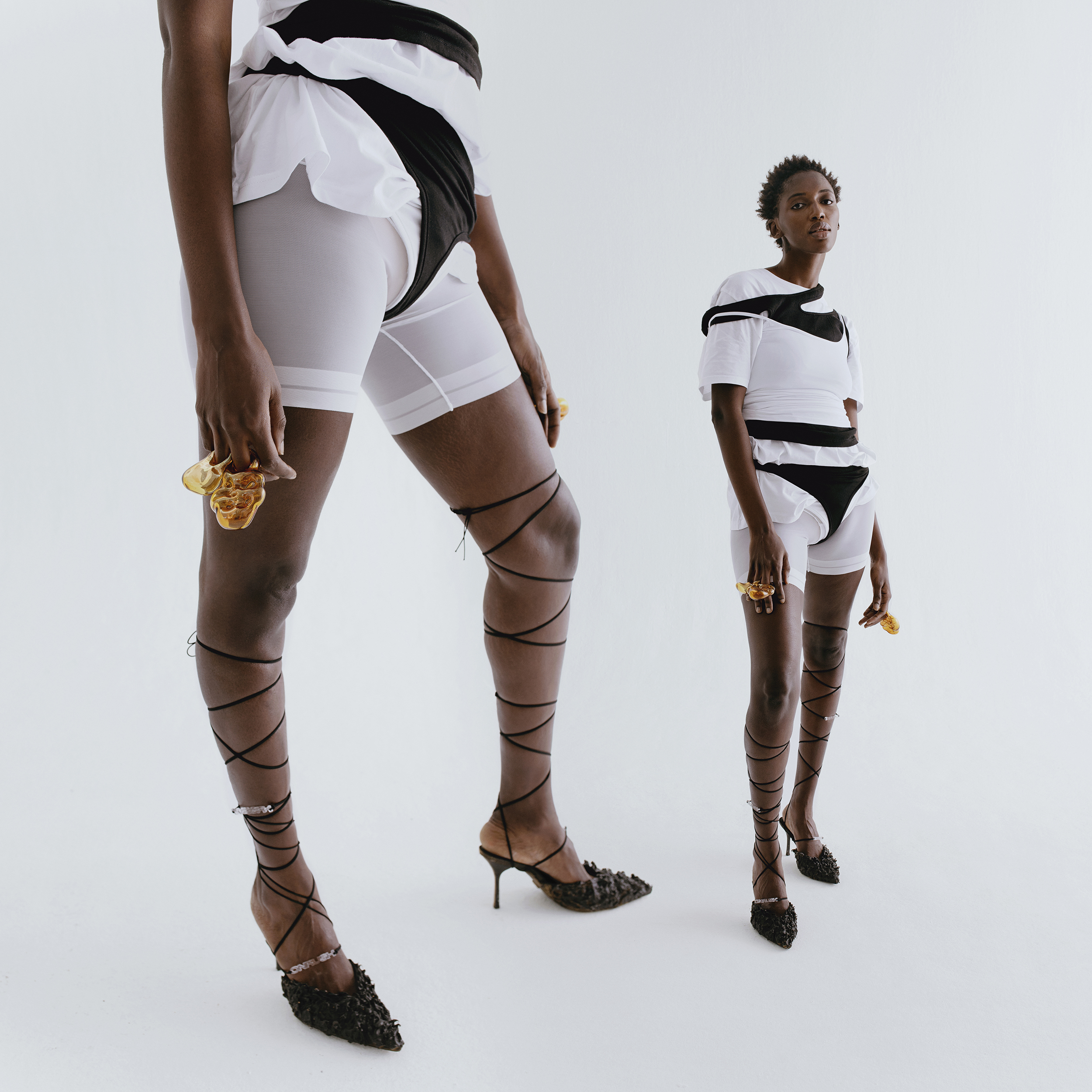 Two dark-skinned adult females pose in white shorts, tops, and strappy heeled shoes against a bright, neutral background in a fashion shot.