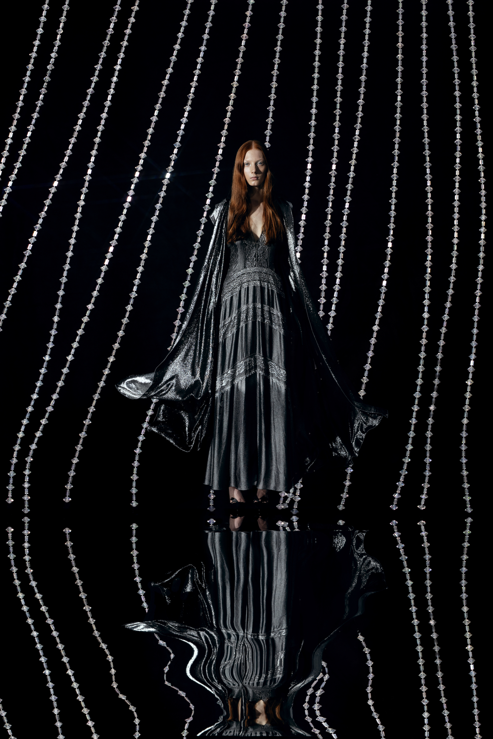 A stylish woman in a metallic dress and cape stands against a backdrop of silver beaded strands in a studio shot, fashion photography.