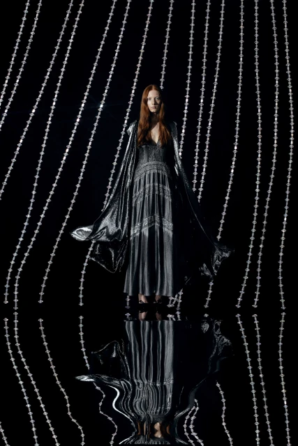 A stylish woman in a metallic dress and cape stands against a backdrop of silver beaded strands in a studio shot, fashion photography.