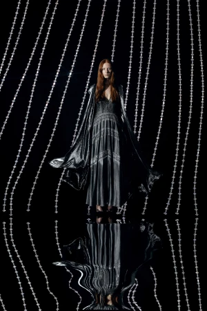 A stylish woman in a metallic dress and cape stands against a backdrop of silver beaded strands in a studio shot, fashion photography.