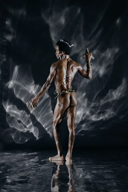 Artistic nude male portrait, rear view, adorned with jewelry and posing against a dark background with textured lighting effects.