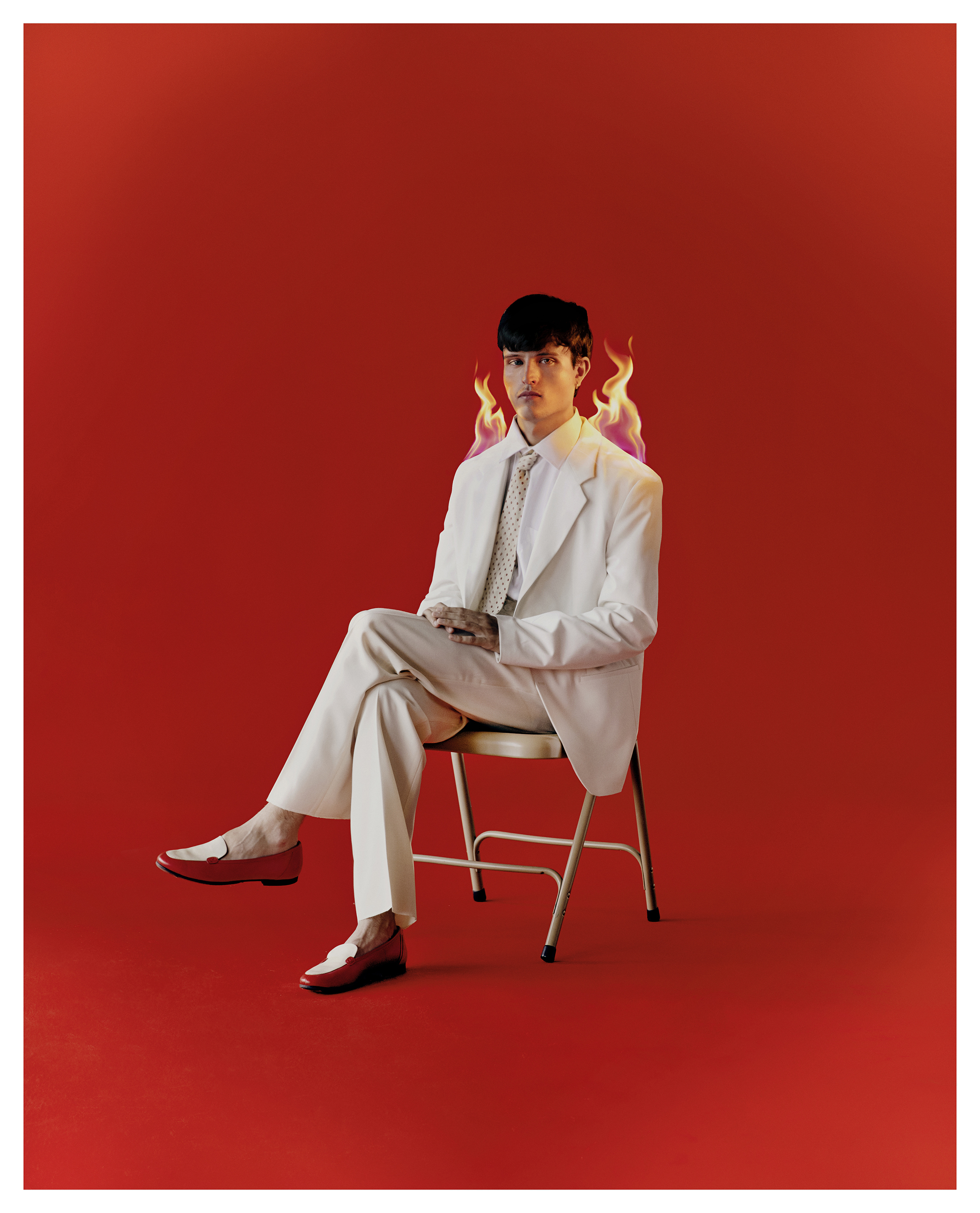 An adult male with short dark hair sits on a chair, wearing a white suit with red loafers, against a vibrant red background with flames.