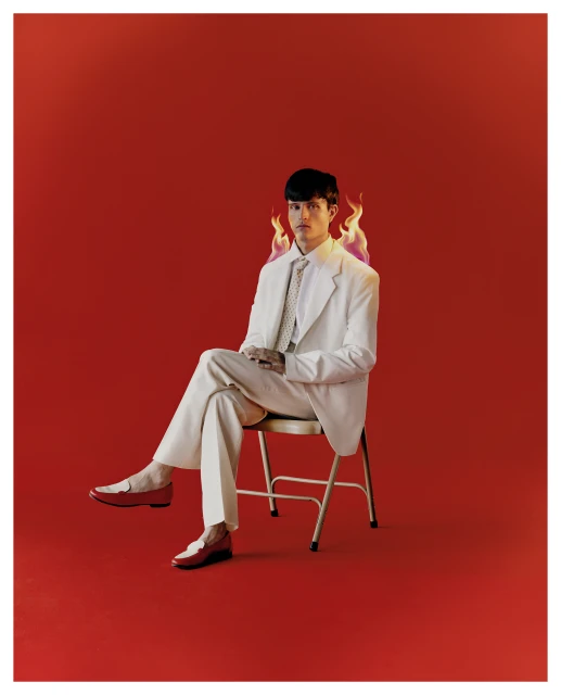 An adult male with short dark hair sits on a chair, wearing a white suit with red loafers, against a vibrant red background with flames.