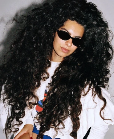 Portrait of a woman with long voluminous dark curly hair wearing black sunglasses and a white t-shirt against a plain background