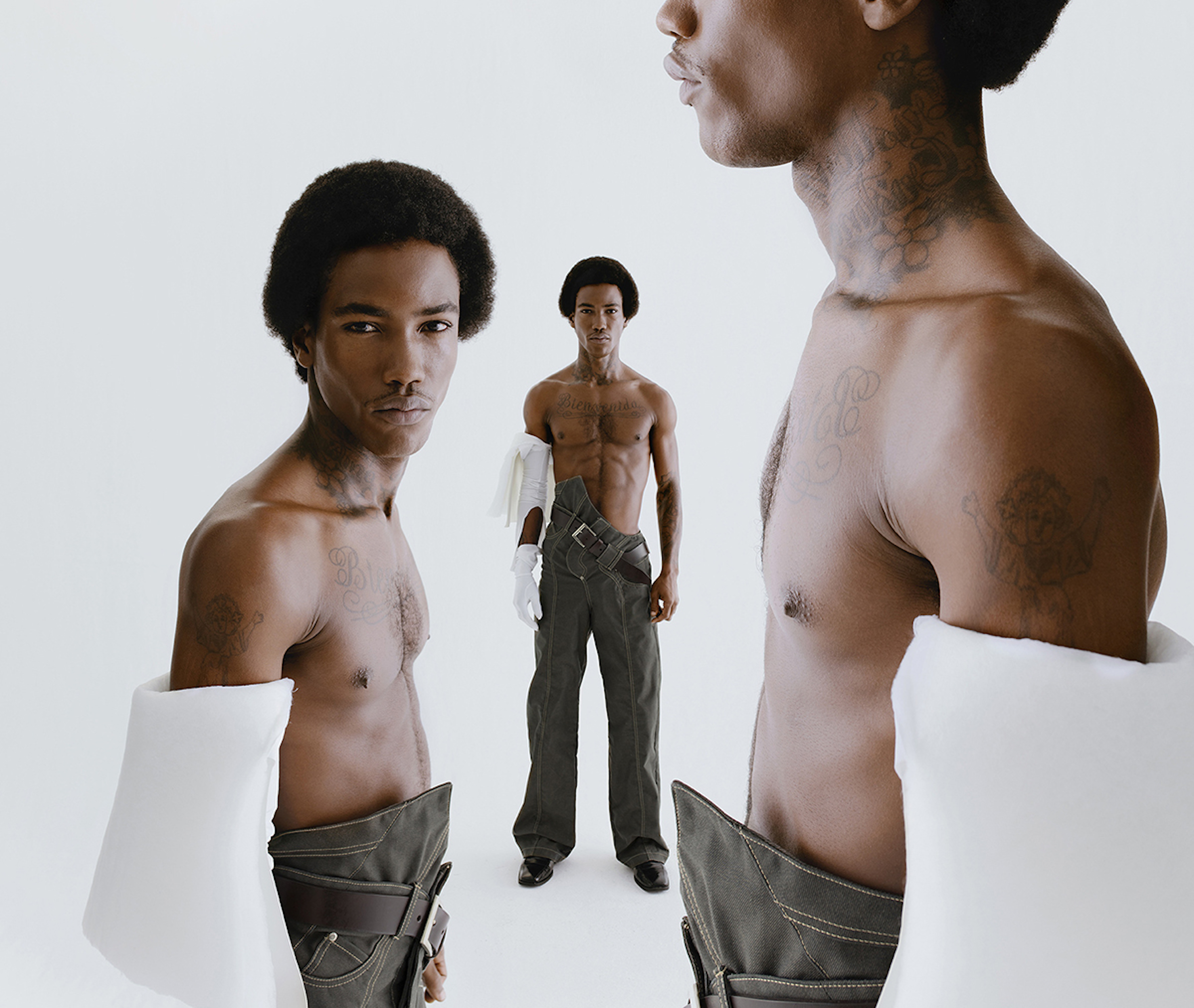 Three African American shirtless men pose in a photo studio with a white backdrop, wearing grey pants, gloves, and arm sleeves.
