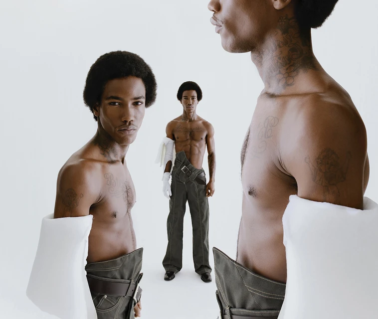 Three African American shirtless men pose in a photo studio with a white backdrop, wearing grey pants, gloves, and arm sleeves.