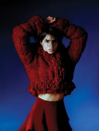 Woman in a textured burgundy coat and skirt posing with arms raised against a moody blue background by Celina Martins