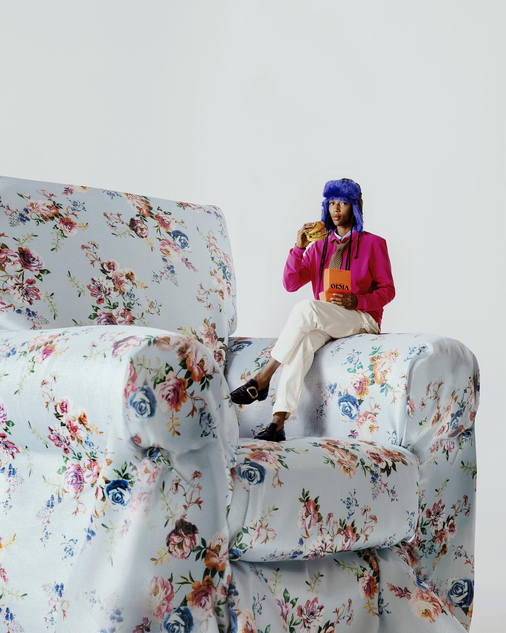 Model wearing a blue fuzzy hat, pink jacket, and white trousers sitting on a massive floral armchair.