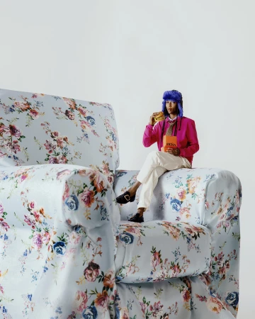 Model wearing a blue fuzzy hat, pink jacket, and white trousers sitting on a massive floral armchair.