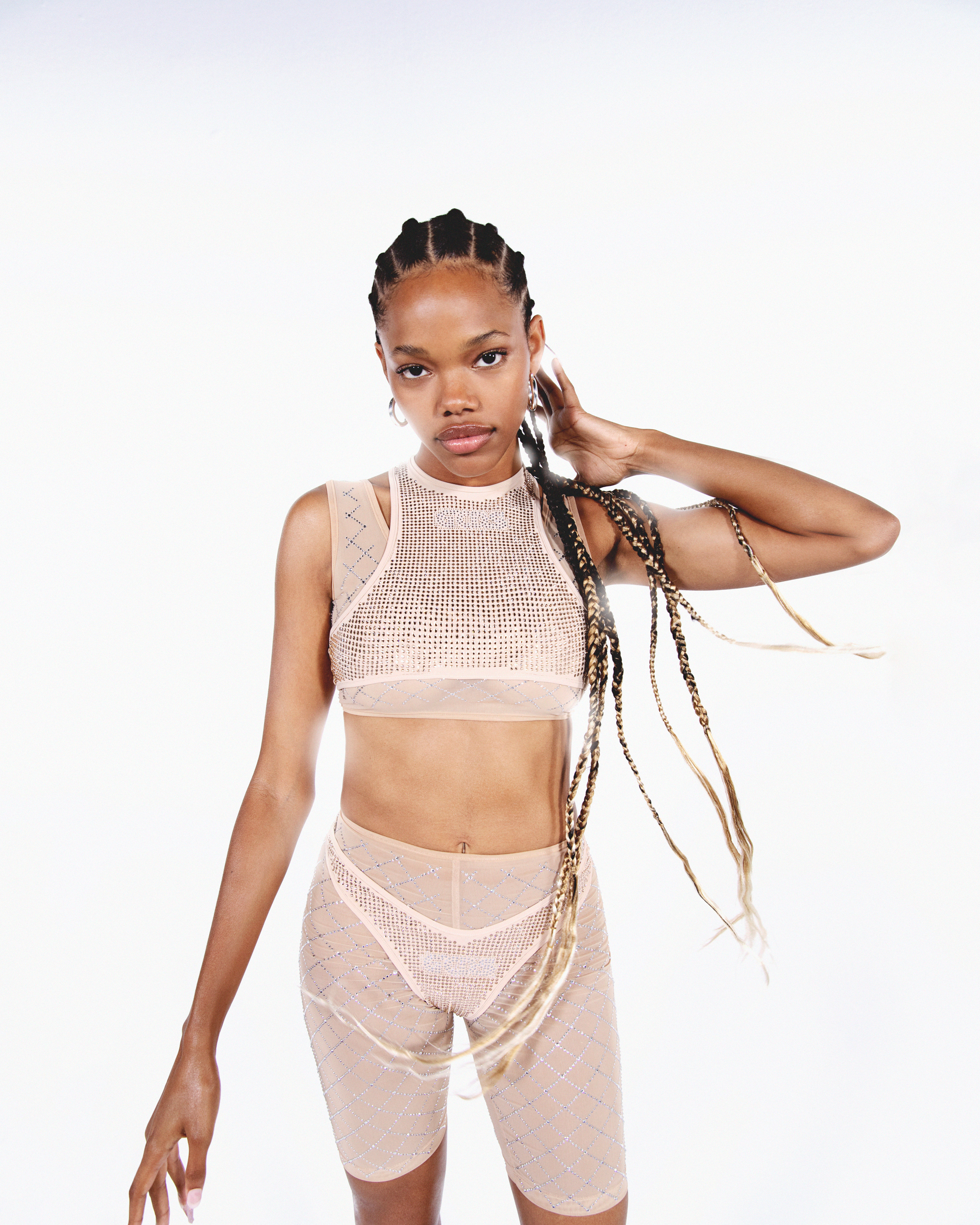 A woman in a stylish two-piece set poses in front of a white background, showcasing her braided hair and neutral-toned athletic wear.