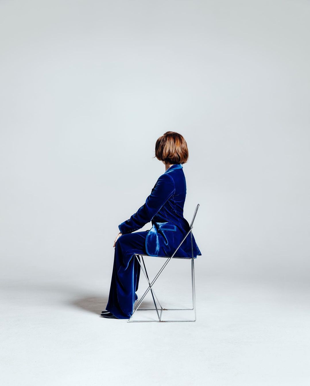 A woman sits on a metal folding chair, facing away from the camera, wearing a vibrant blue velvet suit in a bright photo studio.