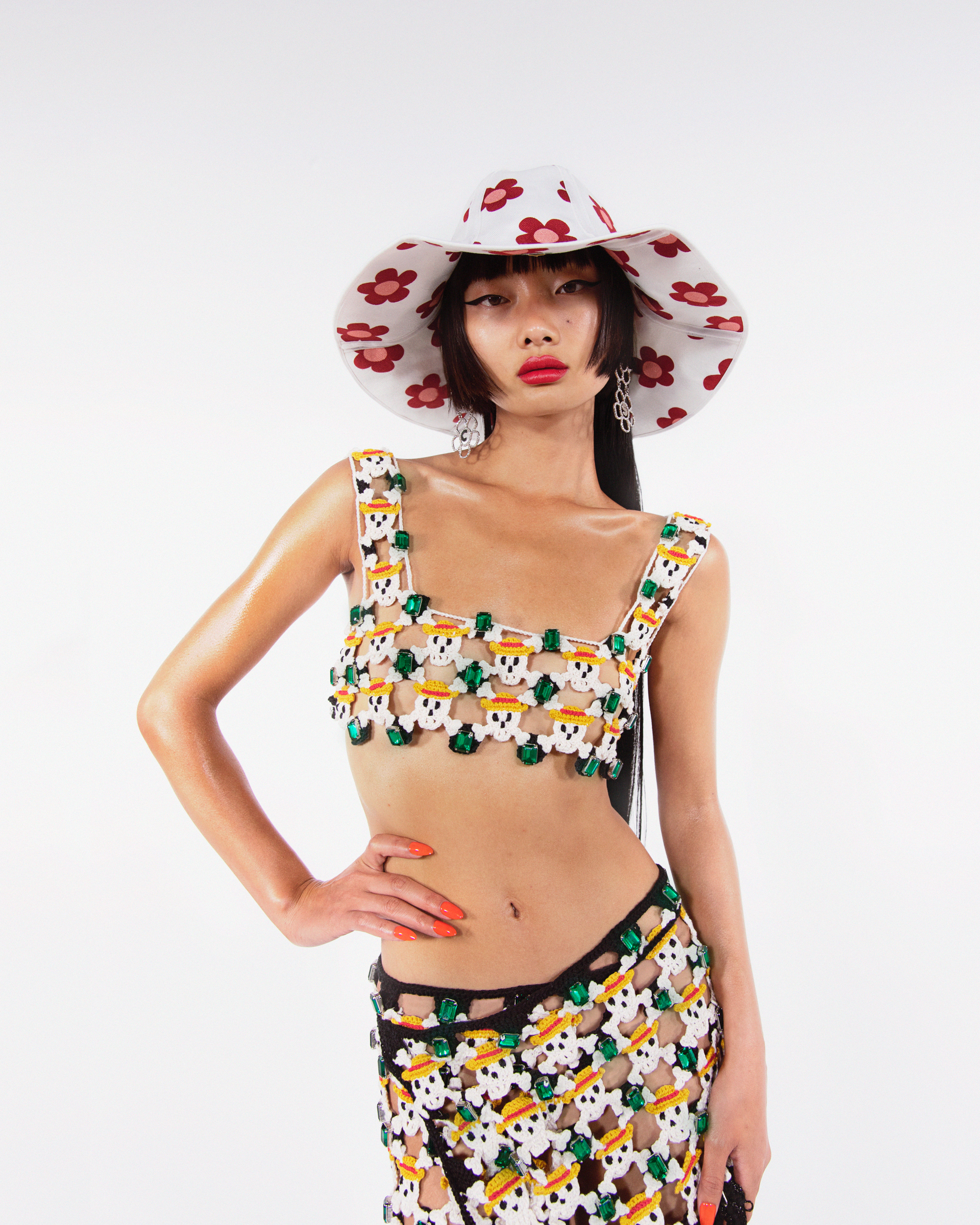 Young Asian woman poses in a white photo studio wearing a hat, top, and skirt made of cartoon skull and gem-studded crochet.