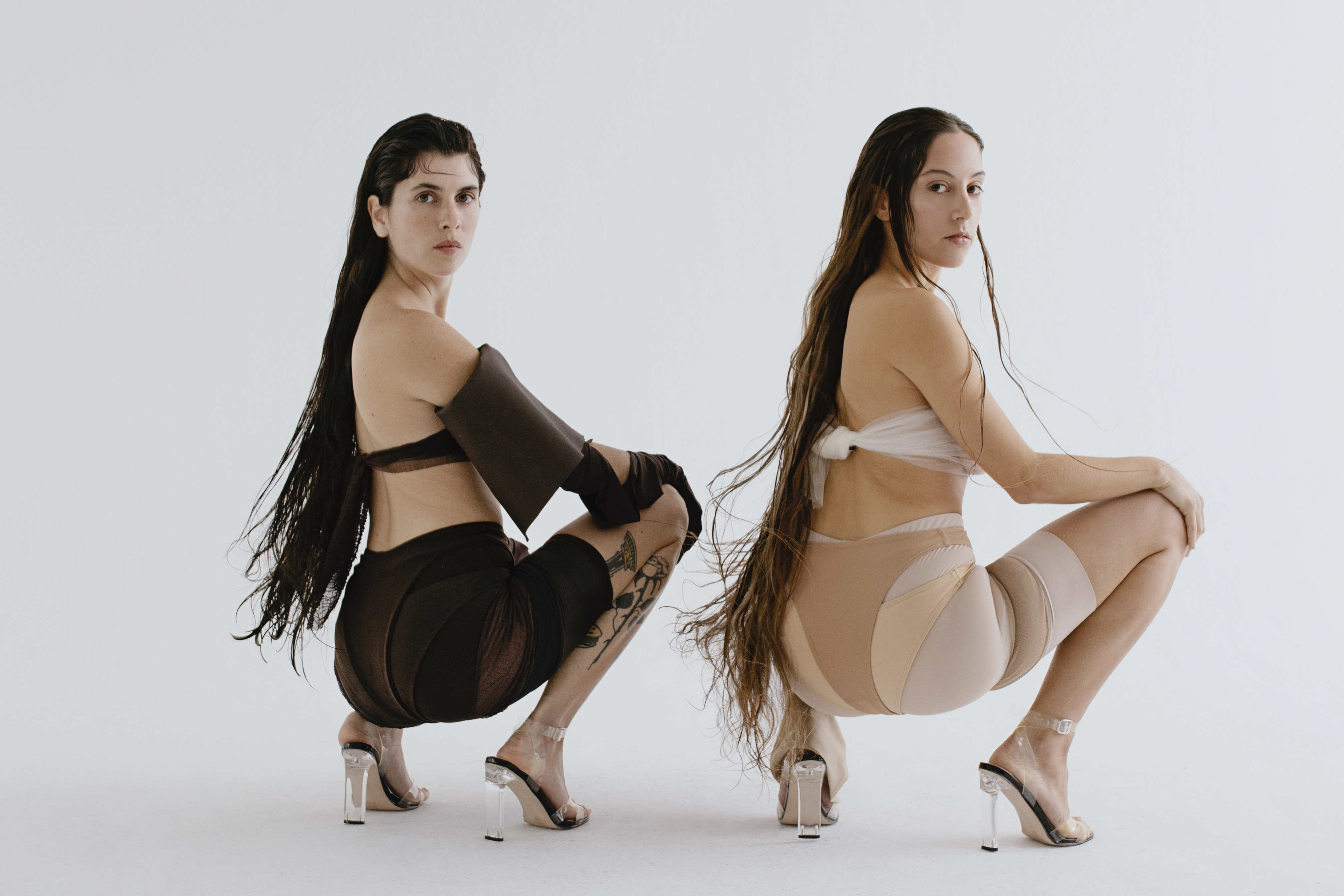Two women in coordinating minimalist fashion pose in a photo studio. They wear heels, with long hair, and neutral makeup against a white background.
