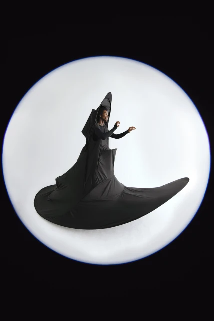 A woman in a black draped gown posing in a studio with a white circular backdrop, captured with a fish-eye lens effect.