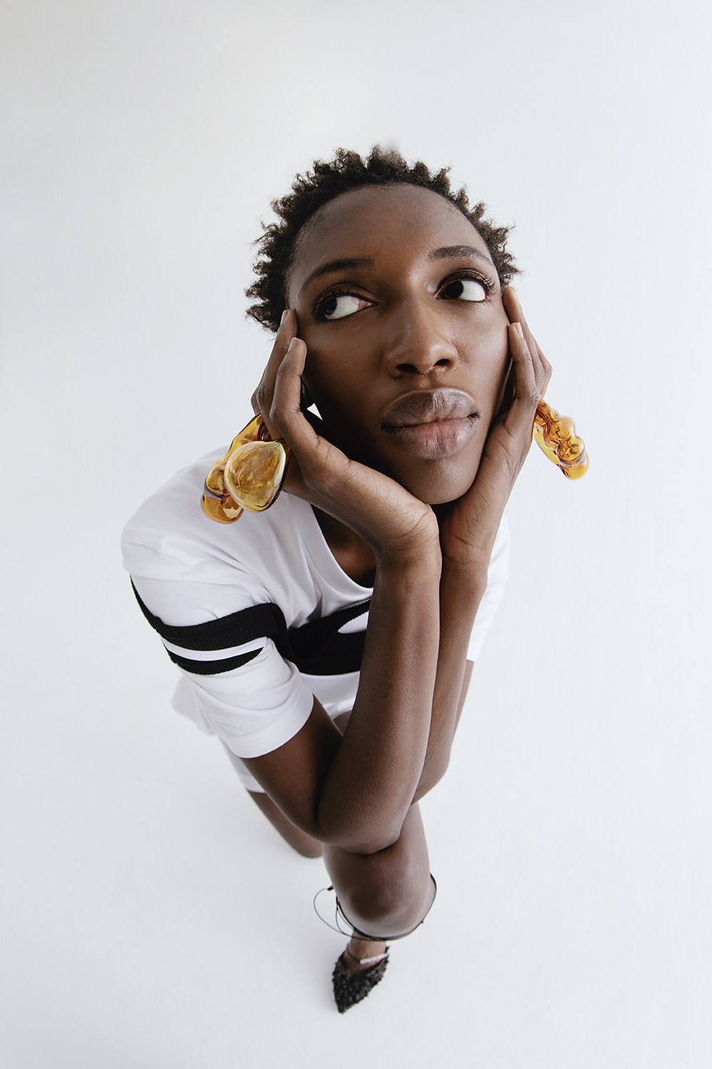 Portrait of a Black woman with short hair, wearing statement earrings and a black and white shirt, looking up against a white background.