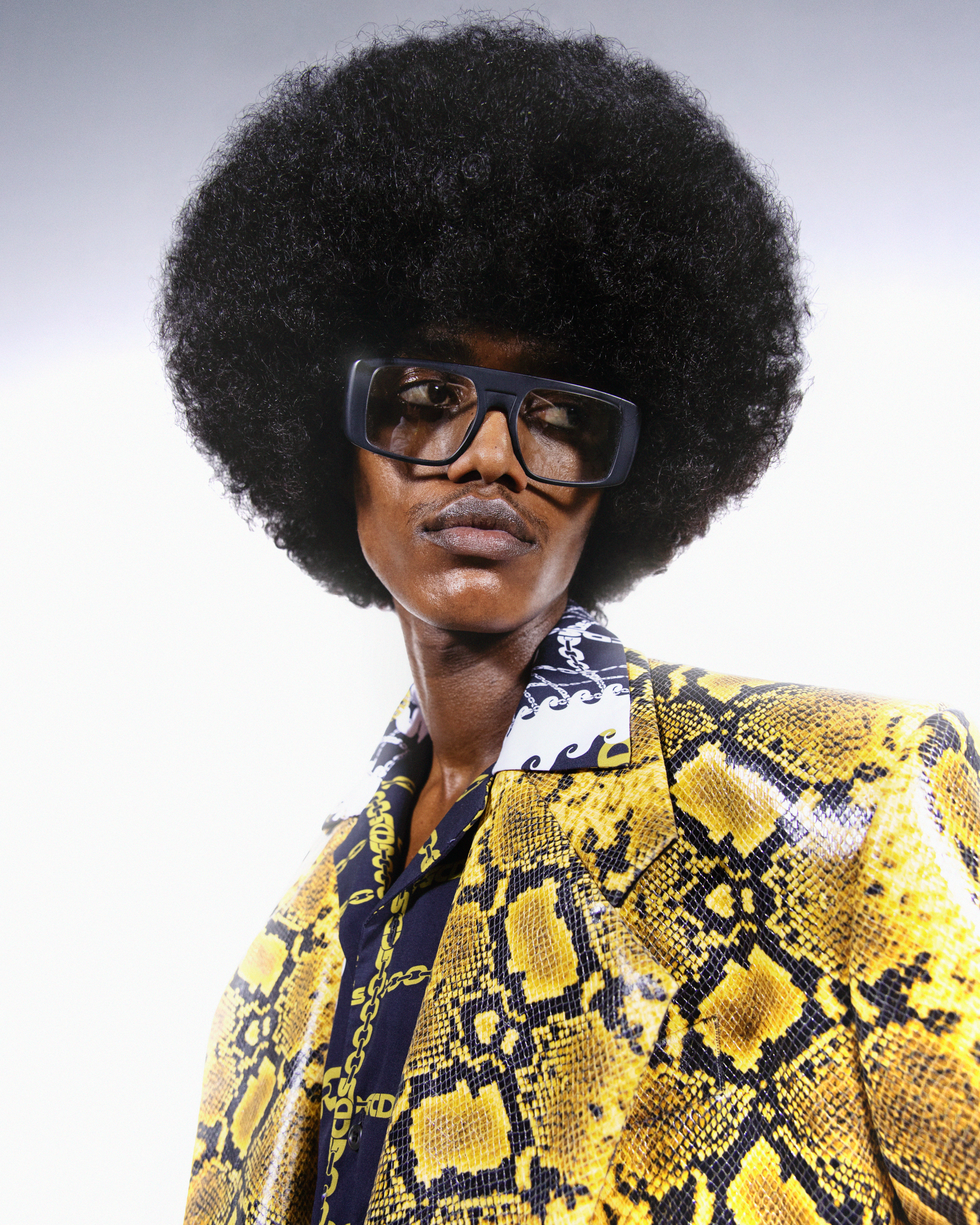 Portrait of a Black male model with an afro hairstyle wearing glasses and a yellow snake-print jacket against a white background.