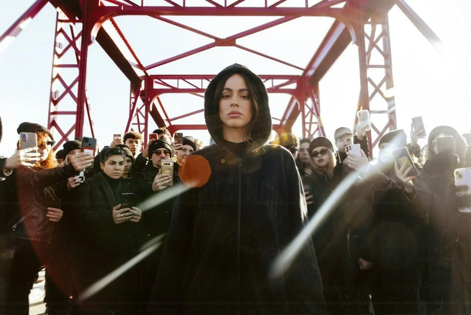 Portrait of a singer in hoodie, standing on a red bridge with a crowd of people taking photos of her in the background.