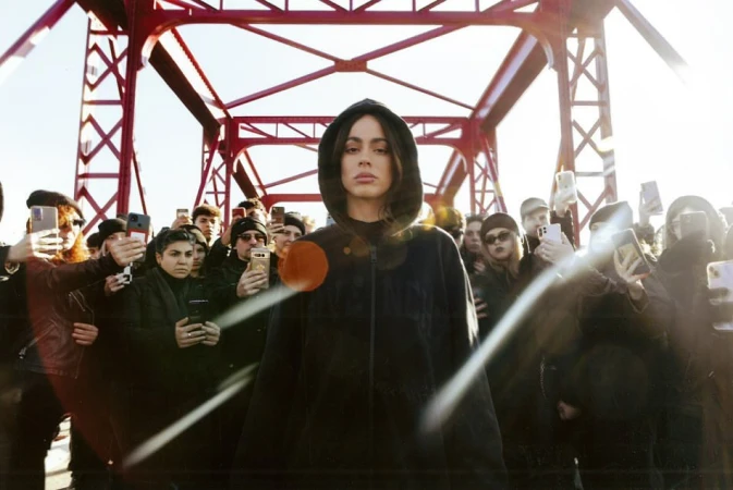 Portrait of a singer in hoodie, standing on a red bridge with a crowd of people taking photos of her in the background.