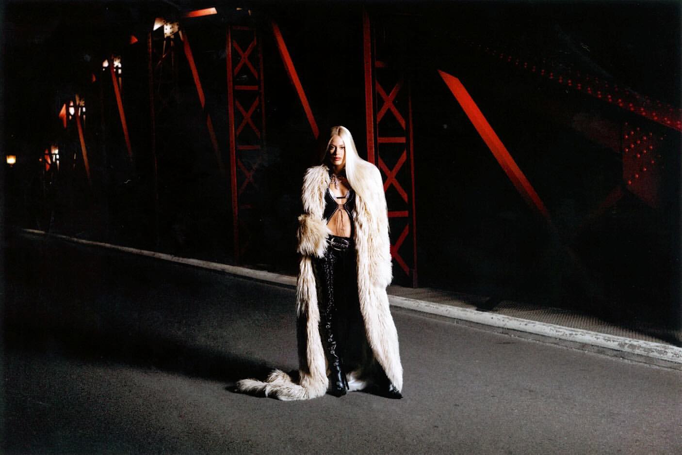 Adult female in a fur coat standing on a bridge at night, illuminated by low lighting with steel beams in the background.